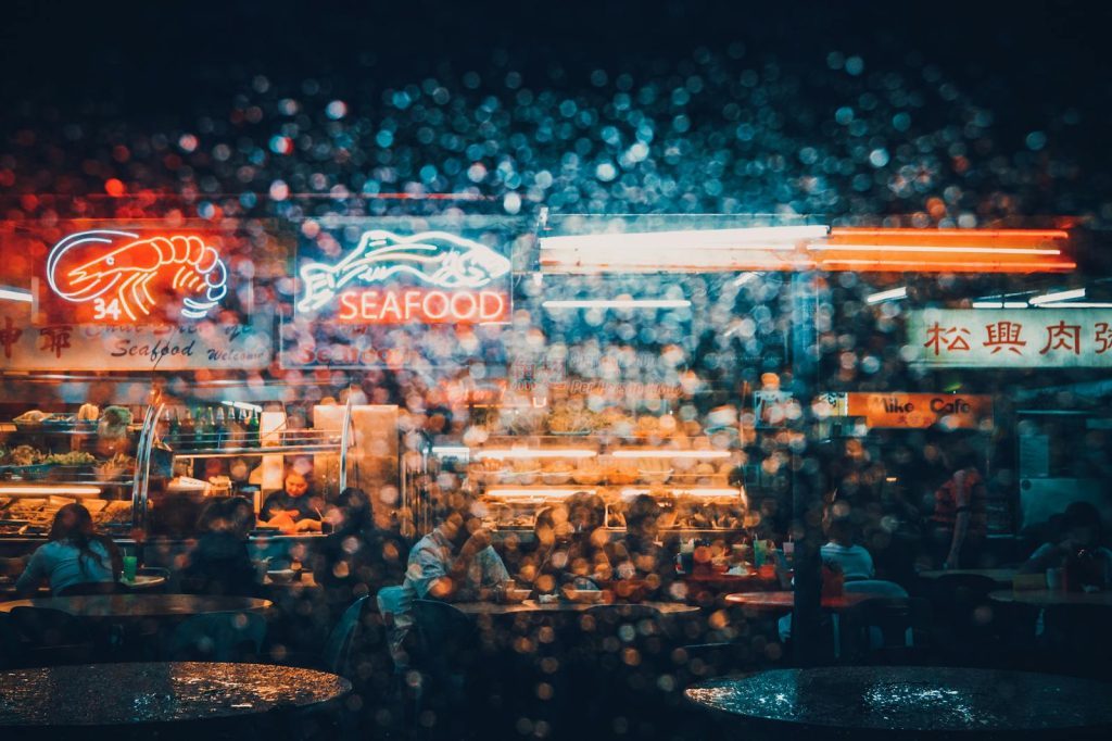 Explore the neon-lit ambiance of a bustling seafood market in Kuching at night, captured through a rain-splattered lens.